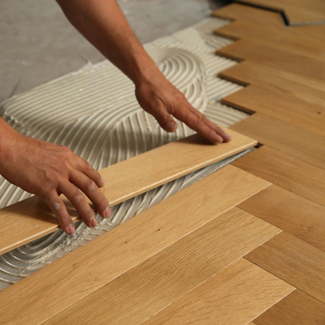 A Fix It Mates technician carefully installing a wooden floor, focusing on precise placement of each plank, with tools and equipment visible in the background, showcasing expert craftsmanship and attention to detail.